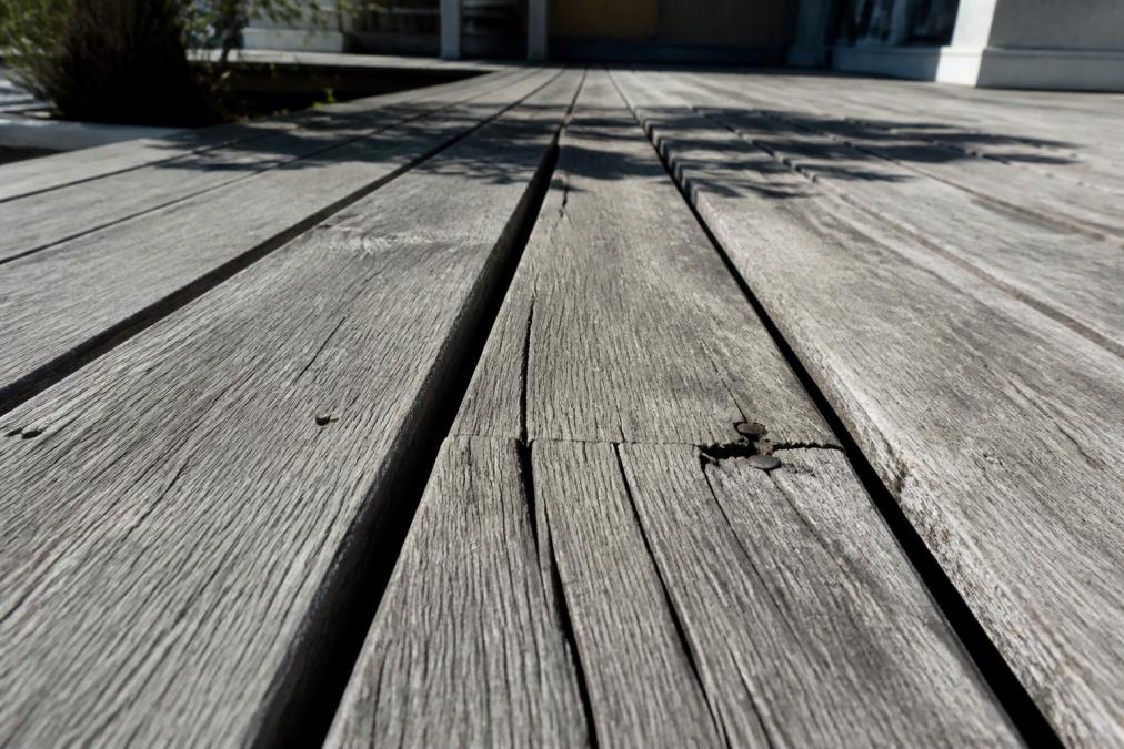 Close-up of weathered, grey timber decking boards with visible cracks, raised splinters and cupped edges under harsh afternoon sunlight
