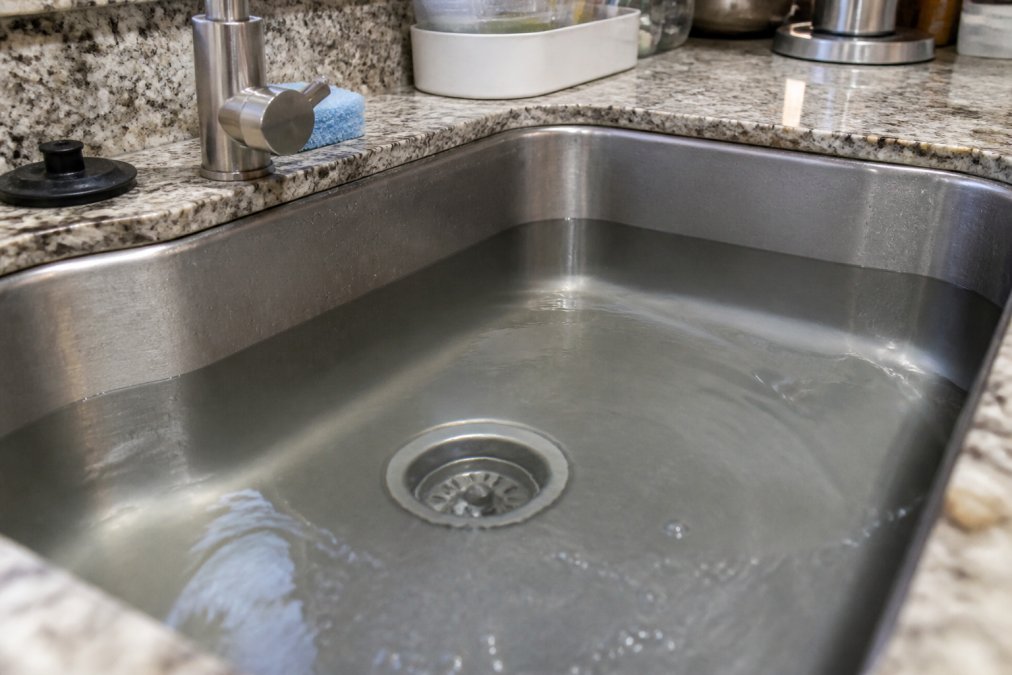 Close-up of water backing up and pooling in a kitchen sink, showing visible water retention and drainage problems that require professional attention, with natural kitchen lighting