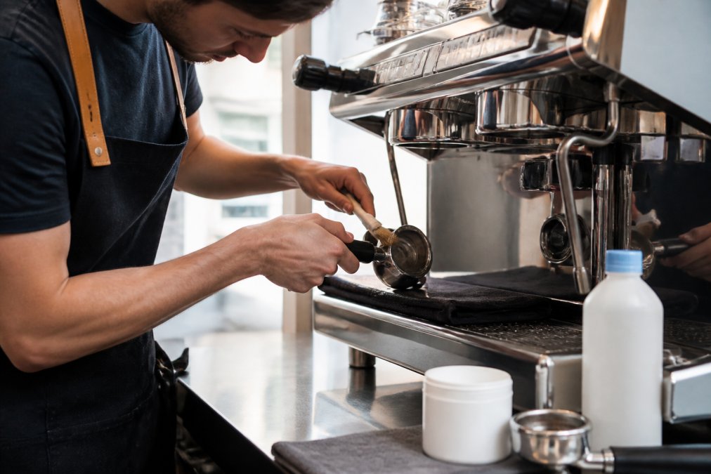 Close-up of hands cleaning the portafilter and group head of an espresso machine with a cleaning brush, cleaning supplies and descaling powder visible on the counter
