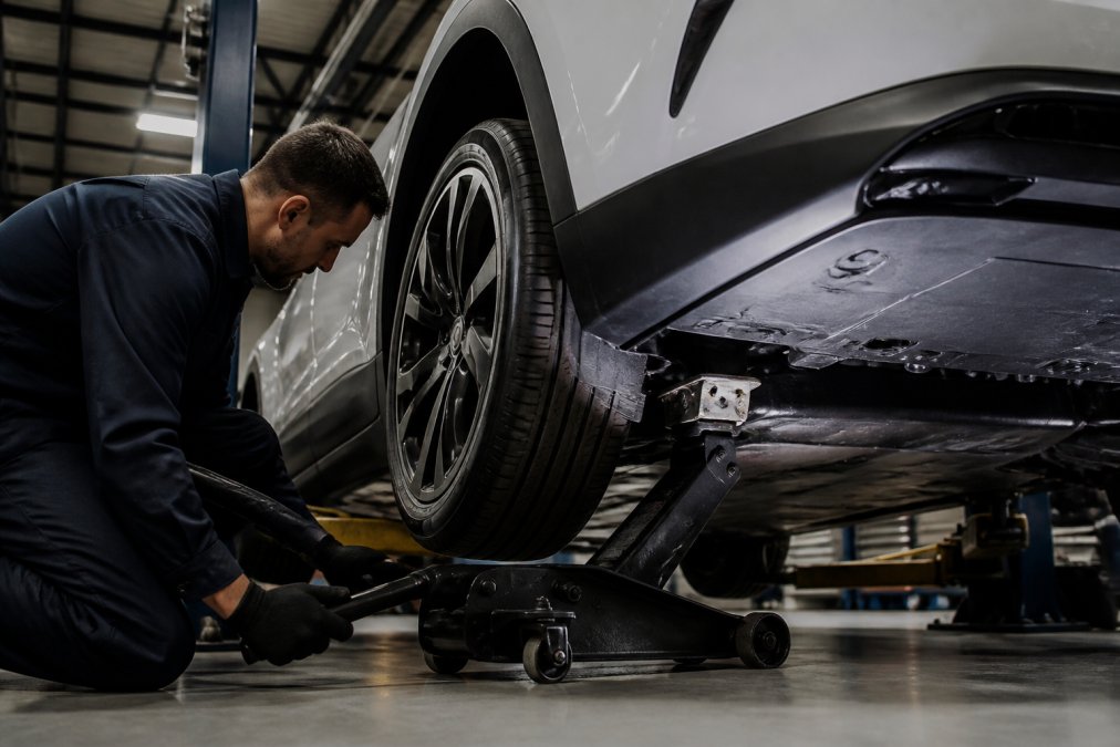 A professional technician in a dark uniform positioning a floor jack under the reinforced lift point of a modern electric sedan inside a clean, well-lit garage, with the battery pack visible underneath the chassis