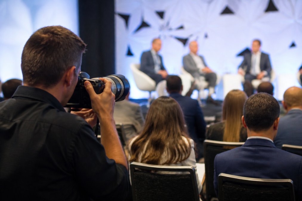 Professional photographer positioned discreetly at the edge of a corporate summit networking reception, capturing genuine conversation between three attendees while maintaining respectful distance and natural lighting, attendees unaware of being photographed