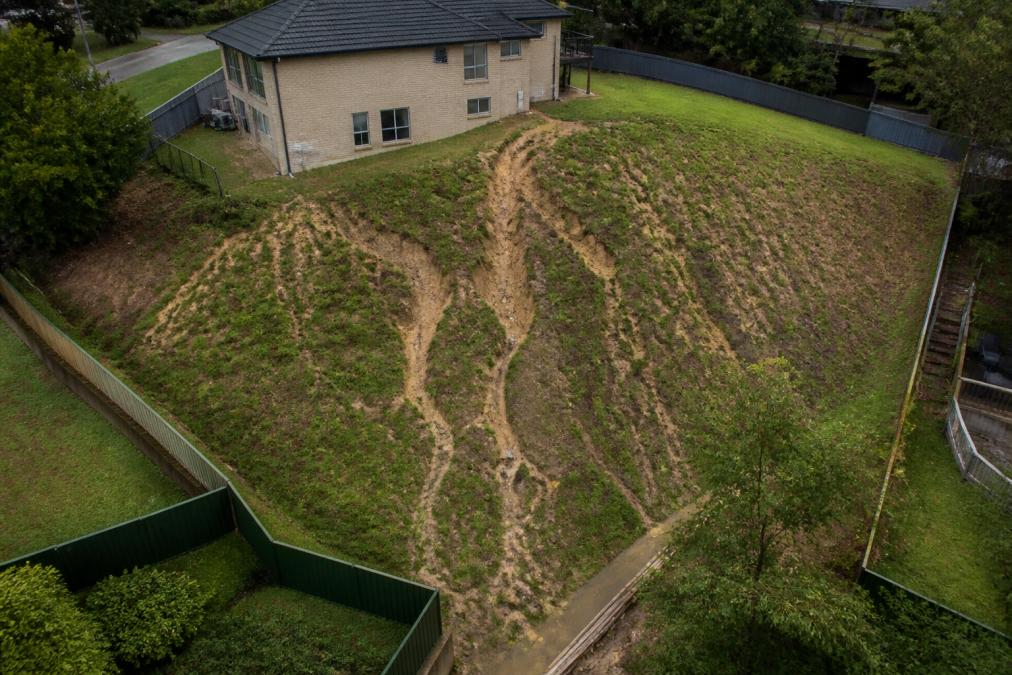 Aerial view of a sloped residential property with uneven terrain, exposed soil patches, and visible water runoff patterns flowing down the hillside in natural daylight