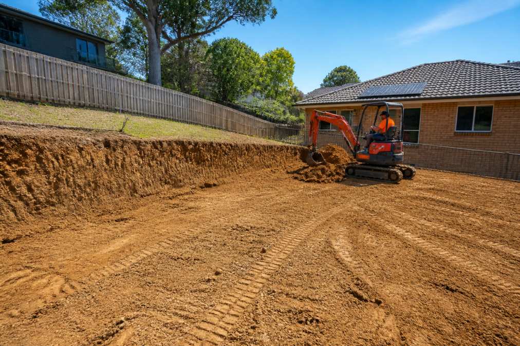 Sloped residential backyard during active excavation with heavy machinery operator managing cut and fill operations, showing graded and compacted earth surface ready for landscaping in daylight