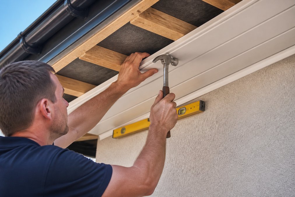 Professional tradesperson installing soffit boards under the eaves, showing proper alignment and fastening with stainless steel nails, close-up of hands positioning board onto battens with spirit level visible