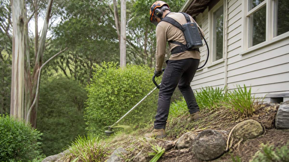 Operator using a battery-powered line trimmer for grounds keeping on a sloped garden bed near a house