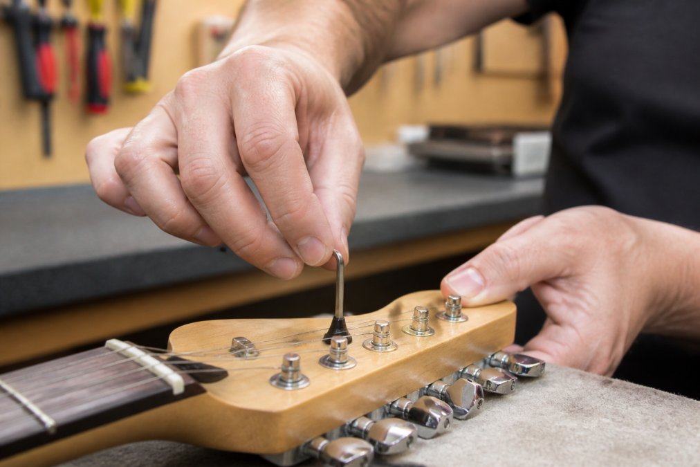 Professional luthier's hands adjusting a truss rod with an Allen wrench on a guitar headstock, showing precise tool placement and careful hand positioning in bright workshop lighting