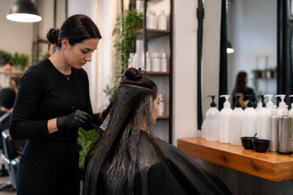 A professional hair stylist applying a smoothing treatment to a client's hair at a salon station, with professional product bottles arranged neatly on the counter, warm salon lighting illuminating the workspace