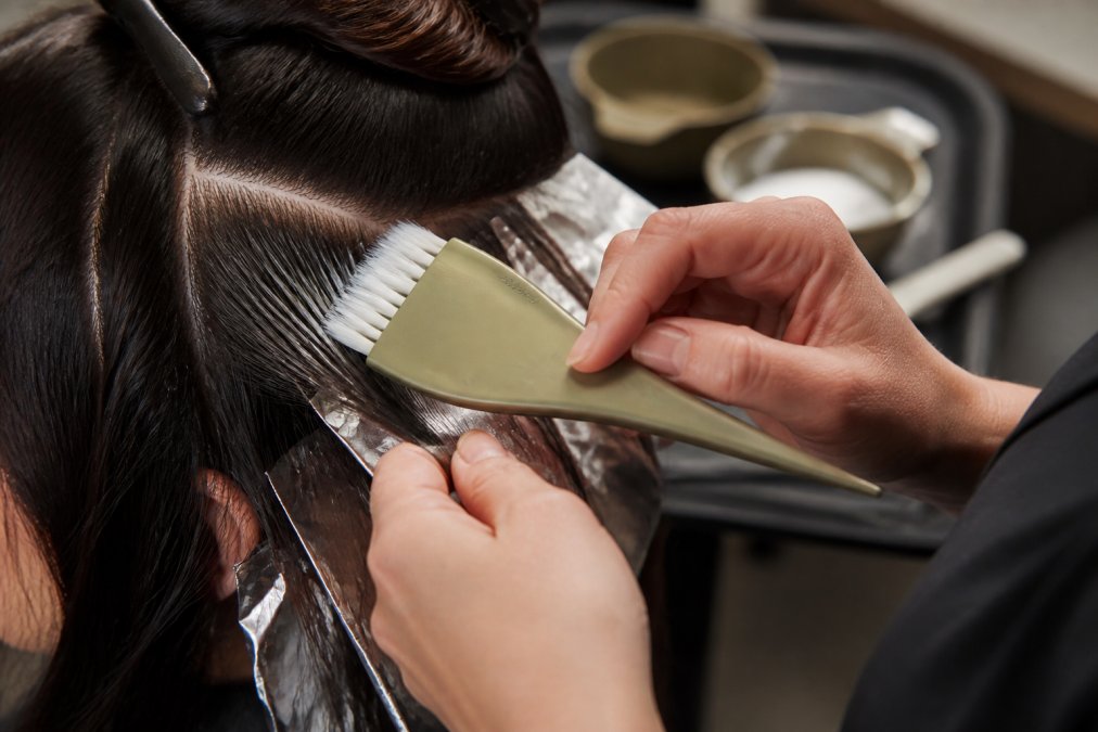 Close-up of a professional colorist's hands applying hair color with a feathered brush to a client's hair, showing precise sectioning and technique at a salon station with color bowls and foils visible