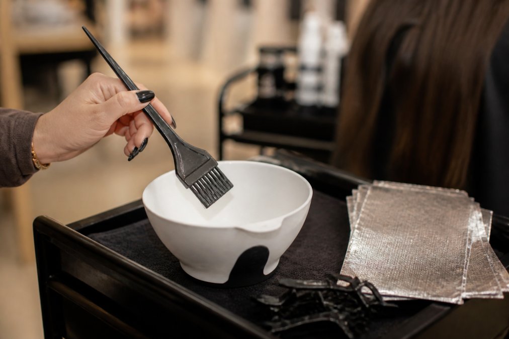 Close-up of a professional colorist's hand holding an ergonomic feathered tint brush over a white stain-resistant mixing bowl, with sectioning clips and highlighting foils visible on the salon station in the soft background, warm salon lighting