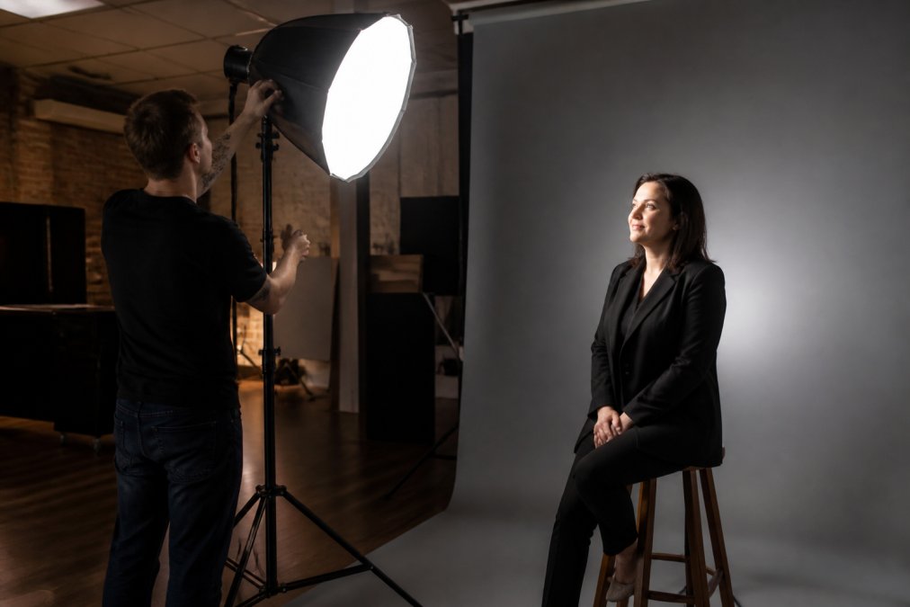 Professional photographer adjusting studio lighting while client sits on stool in front of neutral grey backdrop, bright key light illuminating face