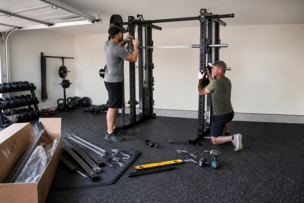 Two people assembling a heavy-duty Smith machine on rubber gym flooring inside a converted garage gym, with tools, a level, and an open equipment box nearby
