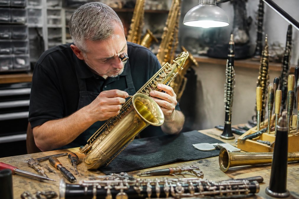 Professional technician examining a saxophone with precision tools, inspecting the keywork and pads under bright workshop lighting, with various brass and woodwind instruments visible on the workbench
