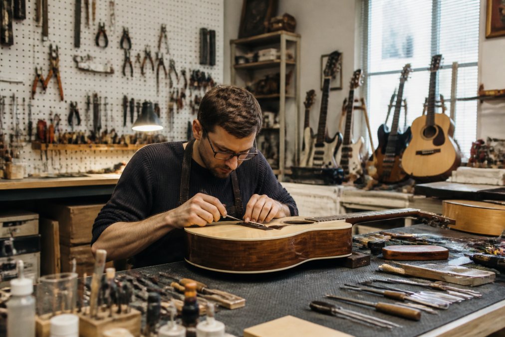 Professional instrument repair workshop with multiple guitars and other instruments on workbenches, specialized tools mounted on pegboards, and a technician carefully working on an acoustic guitar with precision instruments