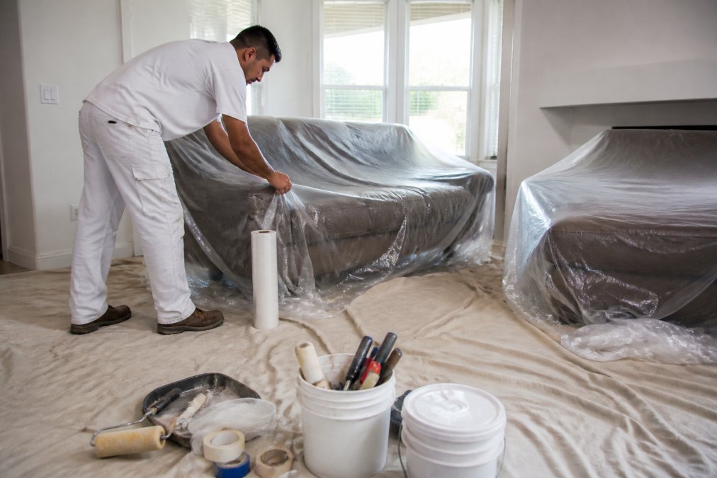 Professional painter applying protective plastic sheeting and drop cloths over furniture and flooring in residential living room with natural light from windows