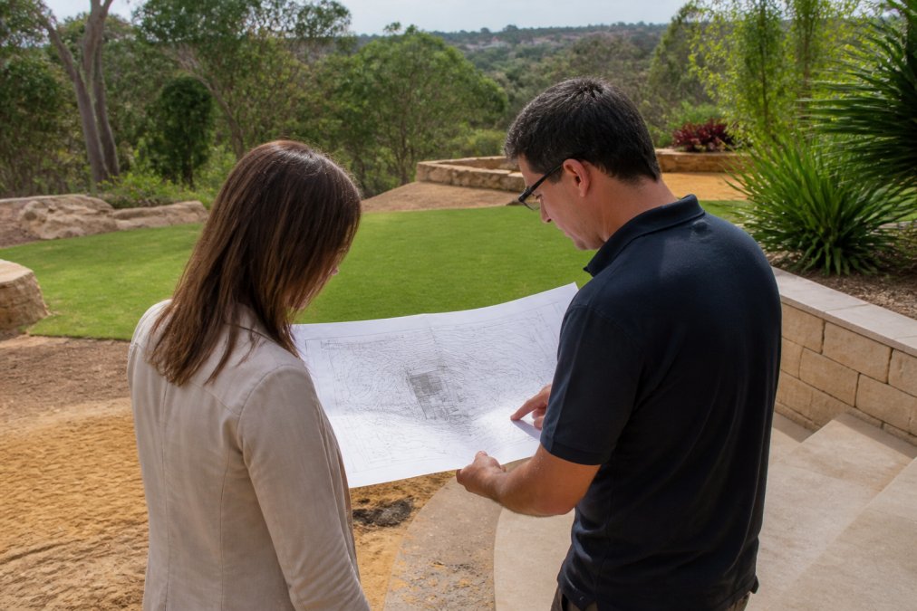 Professional landscape designer and client reviewing site plans and measurements outdoors, with the garden space and topography visible in the background, natural daylight