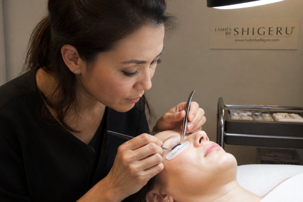 Professional lash technician carefully mapping individual natural lashes on a client's eye using a lash tile and precision tweezers, with bright salon lighting illuminating the delicate work
