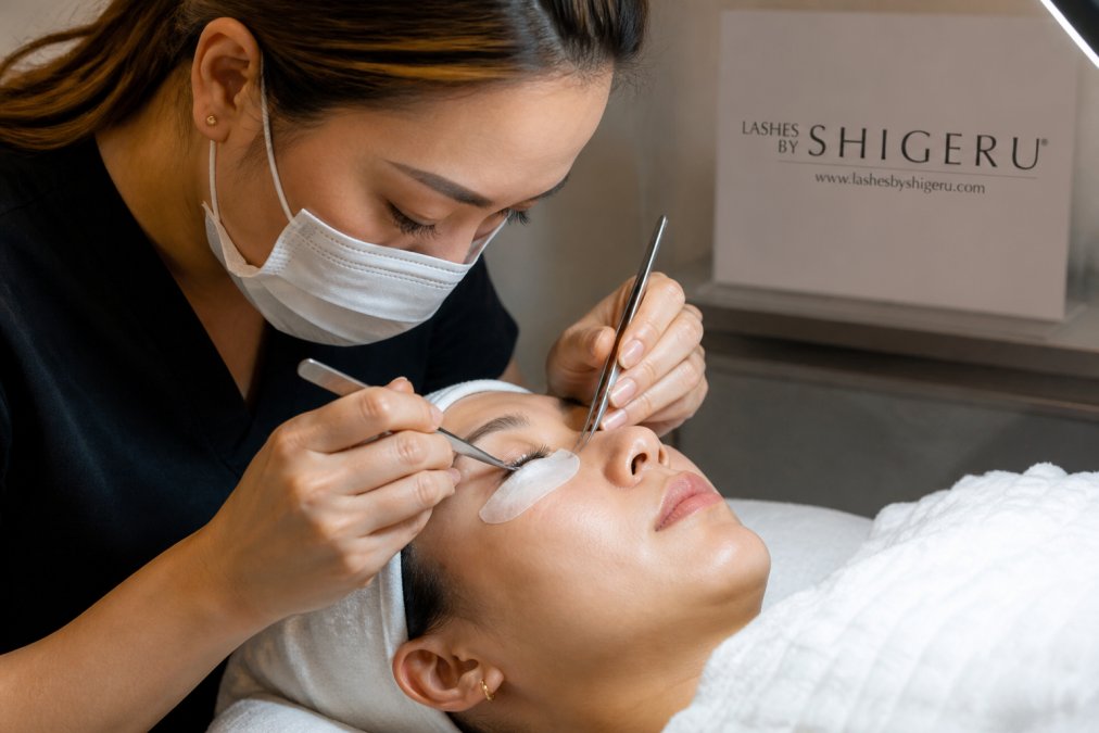 Lash artist's hands using precision tweezers to isolate individual natural eyelashes during the extension application, demonstrating the separation technique under bright professional salon lighting