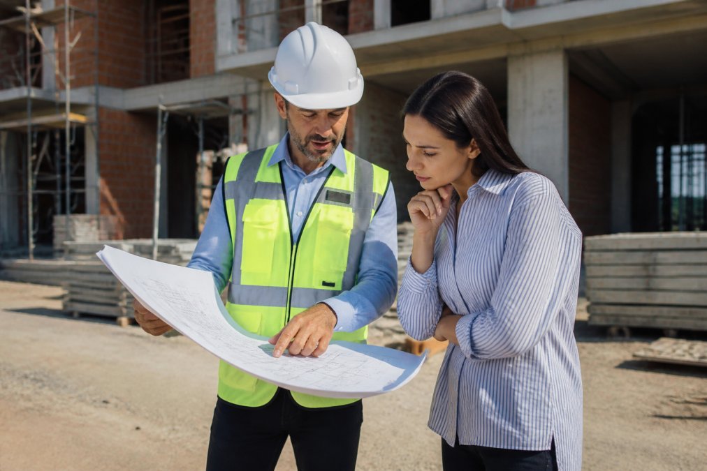 Contractor in hard hat reviewing architectural blueprints with client on construction site, discussing design decisions and workmanship details
