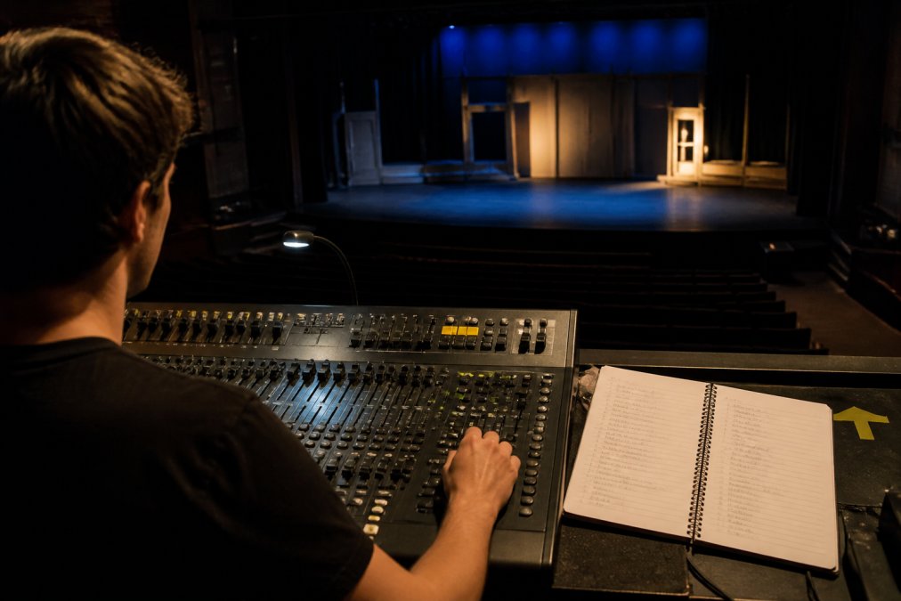 A volunteer lighting operator at a small theater lighting console during a cue run-through, notebook of cue numbers open beside the board, stage visible in the background