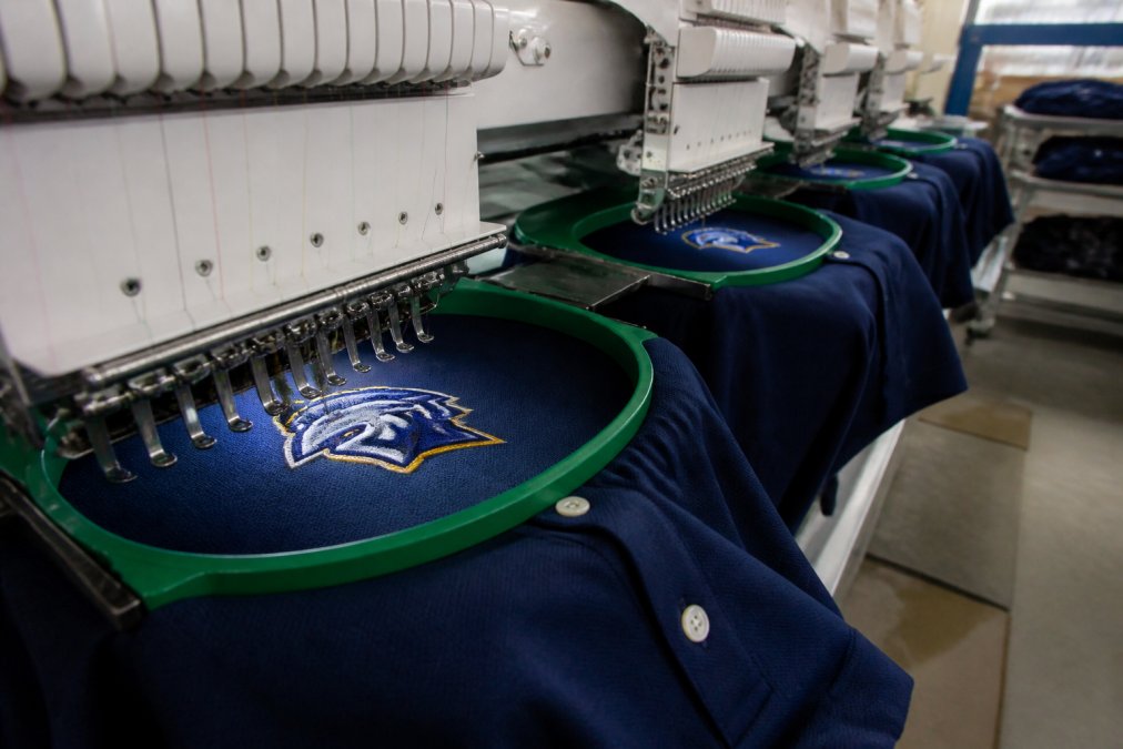 Close-up of a multi-head embroidery machine stitching a team logo onto a navy polo shirt in a bright production studio