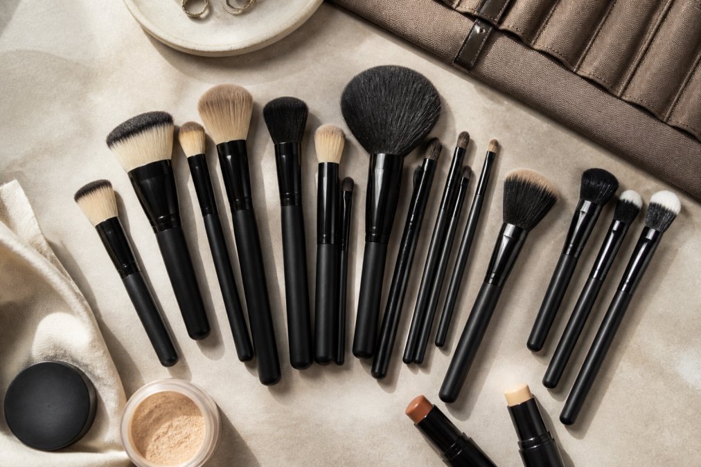 Overhead flat lay of a professional minimal makeup kit arranged on a neutral surface, featuring foundation brushes, lip brushes, and essential application tools with natural lighting