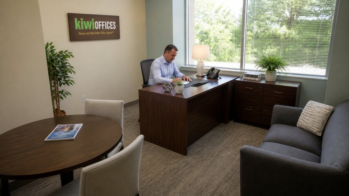 A small business owner working at a desk inside a private furnished office suite, with a client meeting area and natural light from a window