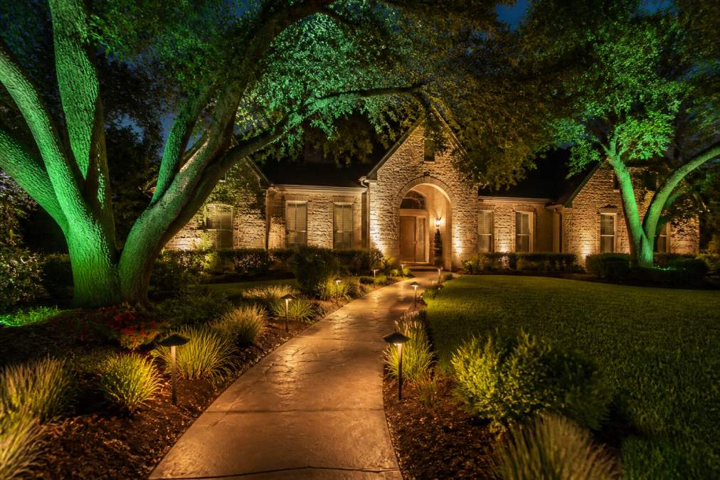 A beautifully lit residential front yard at night showing layered landscape lighting with low path lights lining a curved stone walkway, green uplights on mature oak trees, and soft wash lighting across a brick facade