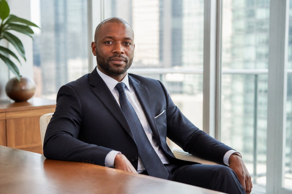 Professional executive in tailored business attire seated in a modern office with floor-to-ceiling windows, natural daylight illuminating the face with warm tones, confident posture, subtle smile