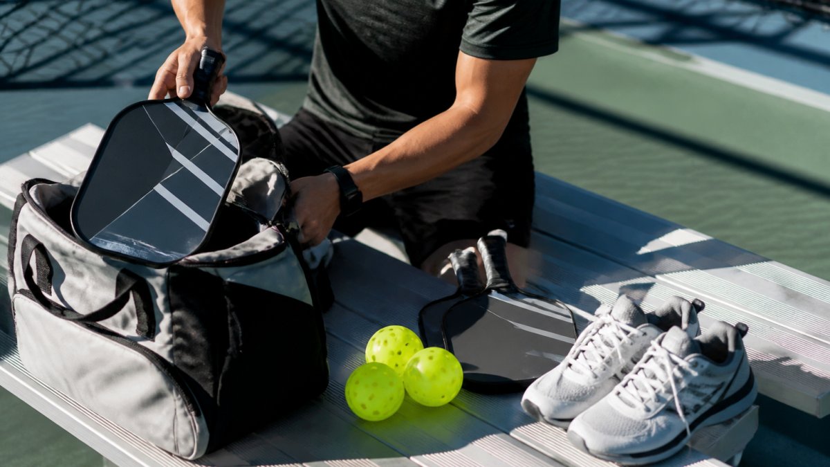 A pickleball player laying out paddles, balls, shoes, and a gear bag on a court bench before a match, natural daylight