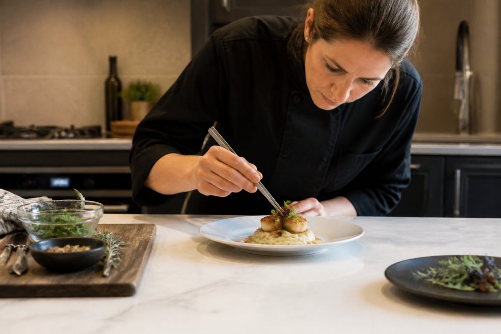 Home cook carefully plating a dish in a residential kitchen, hands positioning food elements with tweezers and precision, warm golden lighting highlighting the finished plate composition