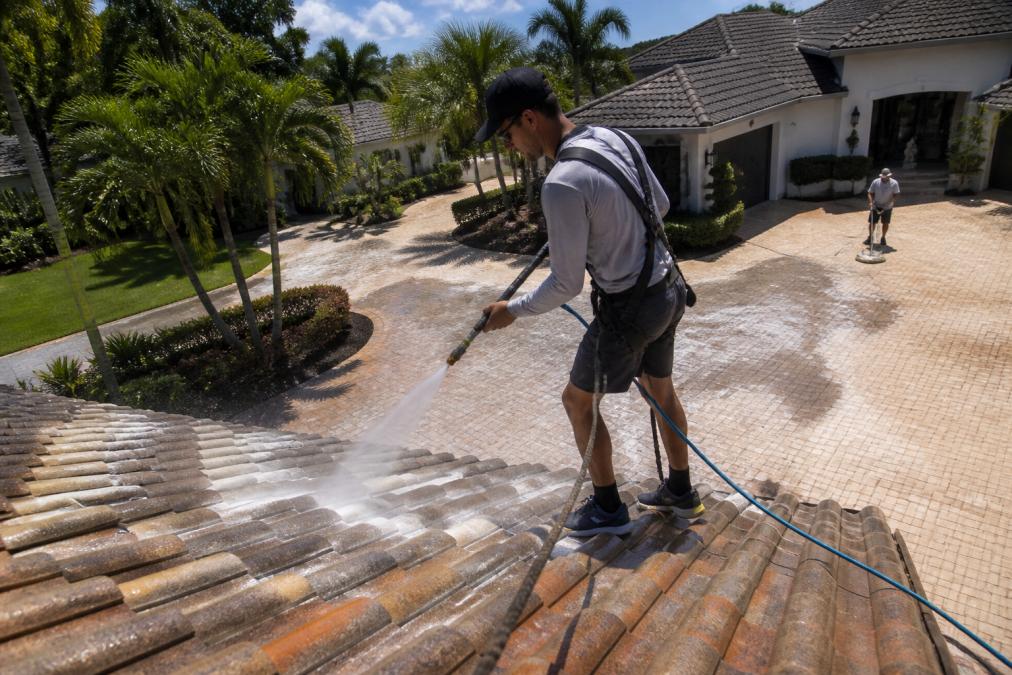 Professional contractor using soft wash equipment on residential roof with safety harness, demonstrating proper technique to safely remove algae and moss without high-pressure damage