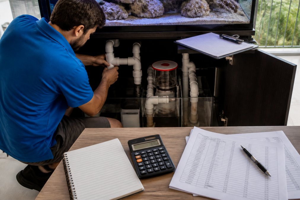 A technician in a blue polo shirt kneeling beside a large reef-ready tank, connecting white PVC plumbing to a sump system while a clipboard with a cost breakdown rests on the cabinet