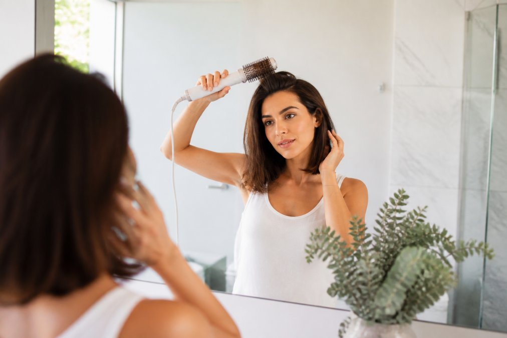 A woman with shoulder-length hair using a white air styler at her roots, lifting the tool upward to create volume, in a bright modern bathroom with natural light