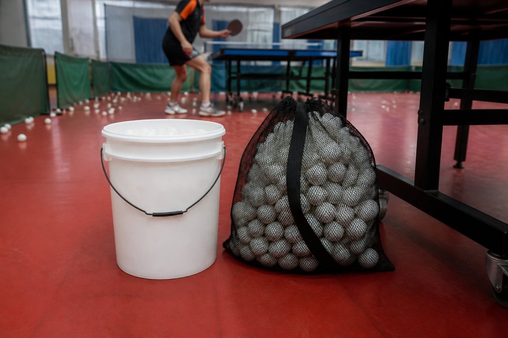 A plastic bucket and a mesh bag filled with white ping pong balls next to a table tennis table leg in a practice hall