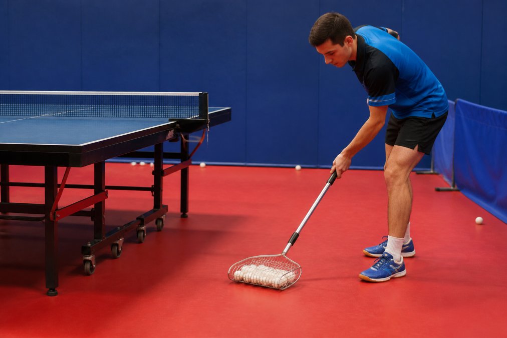 Table tennis player using a ball collection device to scoop up multiple balls from the floor near a practice table, demonstrating ergonomic ease of use and time-saving in action