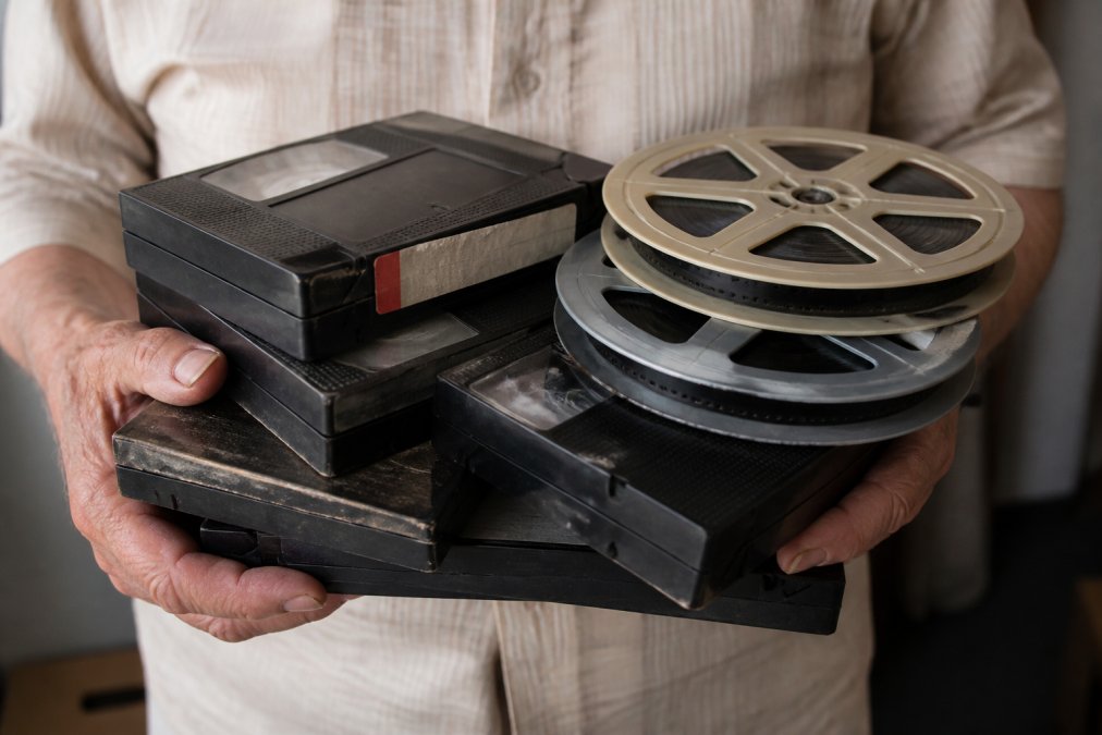 Close-up of weathered hands carefully holding a stack of vintage VHS tapes and 8mm film reels in soft natural light, showing the fragile, aged condition of the media