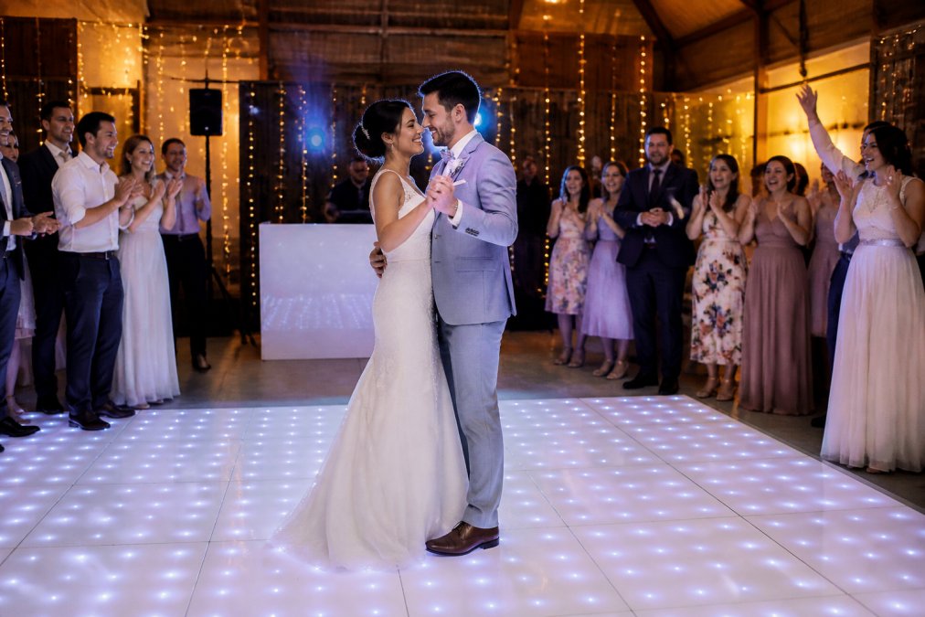 Een bruidspaar danst op een verlichte LED dansvloer tijdens hun bruiloftsfeest, gasten juichen aan de rand van de dansvloer terwijl de DJ in de achtergrond de muziek draait, warme sfeerverlichting in de feestzaal