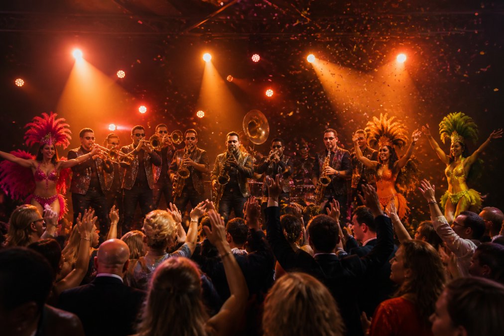 Energieke brassband in volle gang op podium met dansers in felgekleurde outfits, publiek juichend in beweging, warm podiumlicht en confetti in de lucht