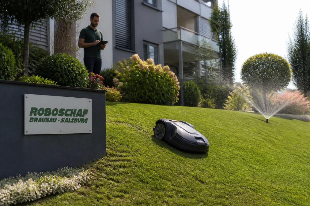 Ein Mähroboter bei der Arbeit auf einer steilen, gepflegten Grünfläche vor einem modernen Wohngebäude, im Hintergrund ein Verwalter mit Tablet, der den Fortschritt beobachtet