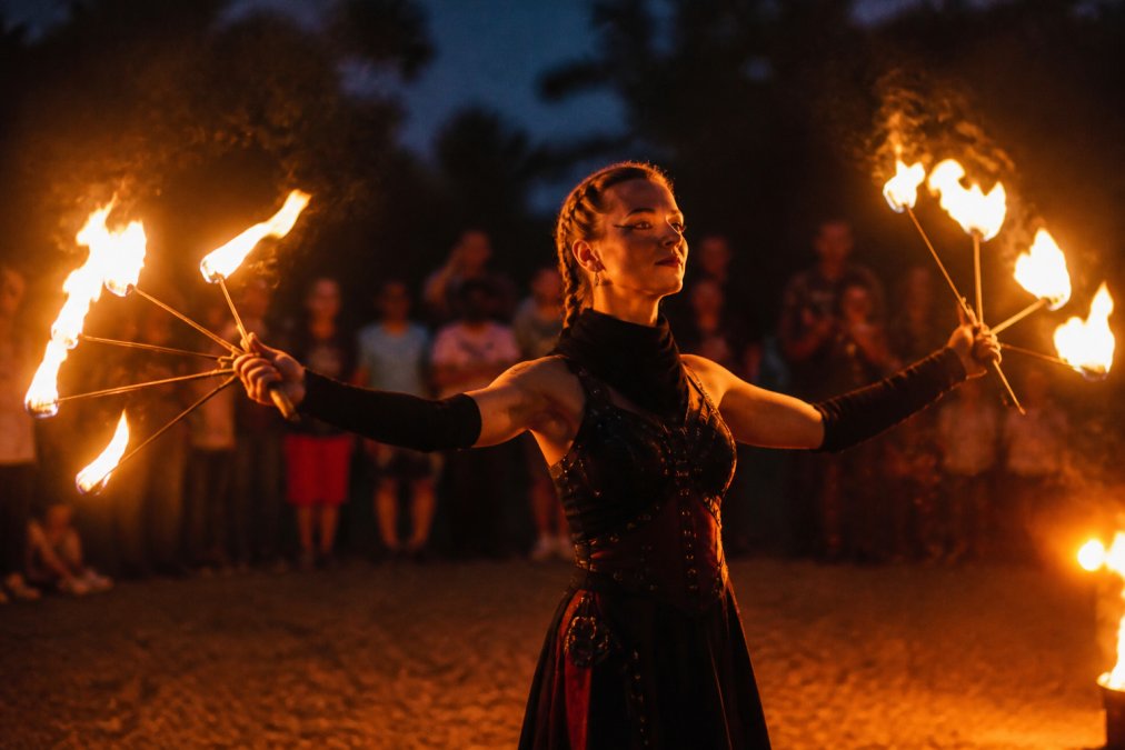 En erfaren ildartist udfører et spektakulært ildshow med dansende flammer og kontrolleret teknik, publikum ses uskarpt i baggrunden