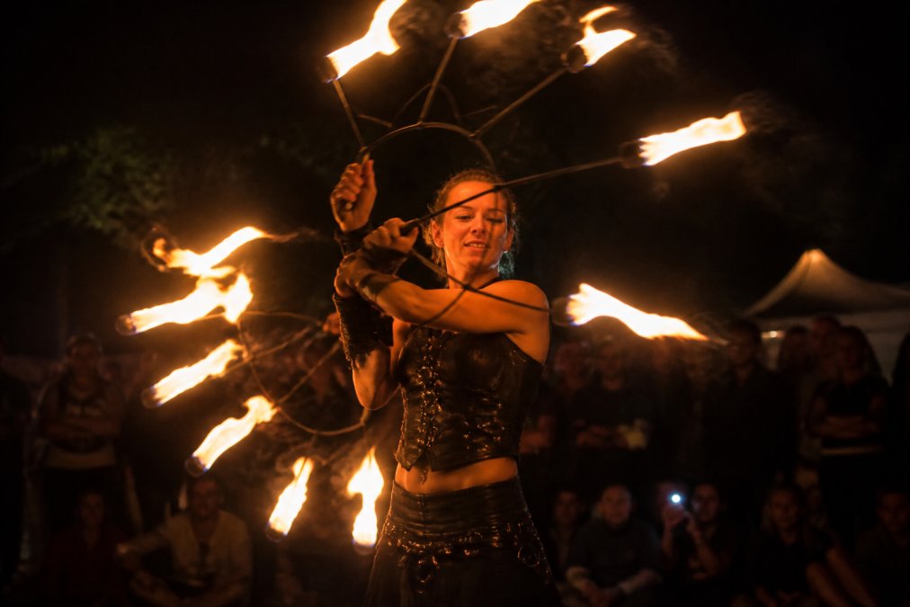 En ildartist optræder med flammende ildhjul i mørket på et udendørs event med publikum i baggrunden, dramatisk belysning fra ilden
