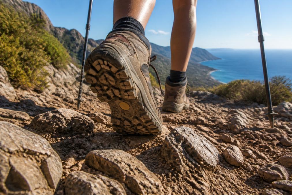 Close-up of hiking shoe soles with deep lugs gripping a rocky, uneven trail in a Mediterranean coastal landscape, with a hiker's legs and trekking poles visible in the background