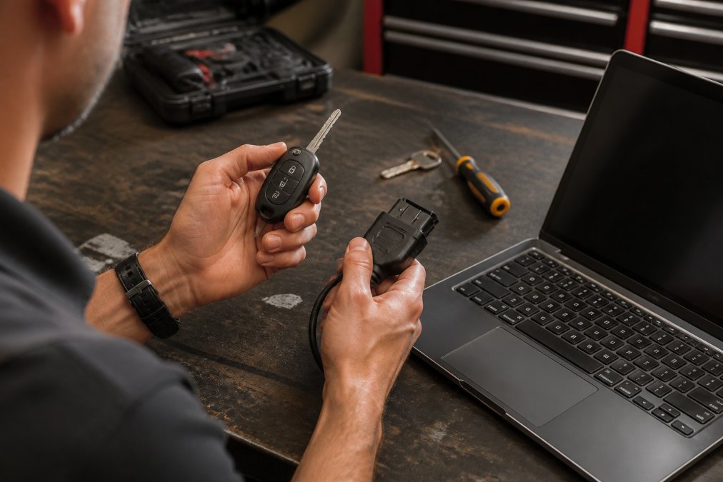 A close-up of hands holding a car key fob and a small OBDII programming tool next to a laptop on a workbench, with a key blank and screwdriver visible in the background