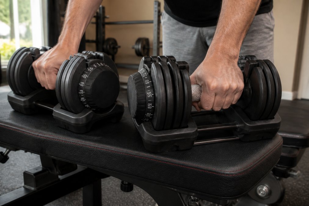 Close-up of hands gripping adjustable dumbbells on a home gym bench, showing progression from lighter to heavier weights with clear weight markings visible