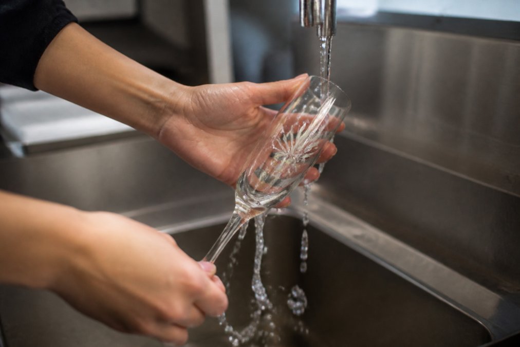 Hands holding a custom-engraved champagne flute being rinsed under running water in a professional kitchen sink, demonstrating everyday durability and commercial dishwasher readiness
