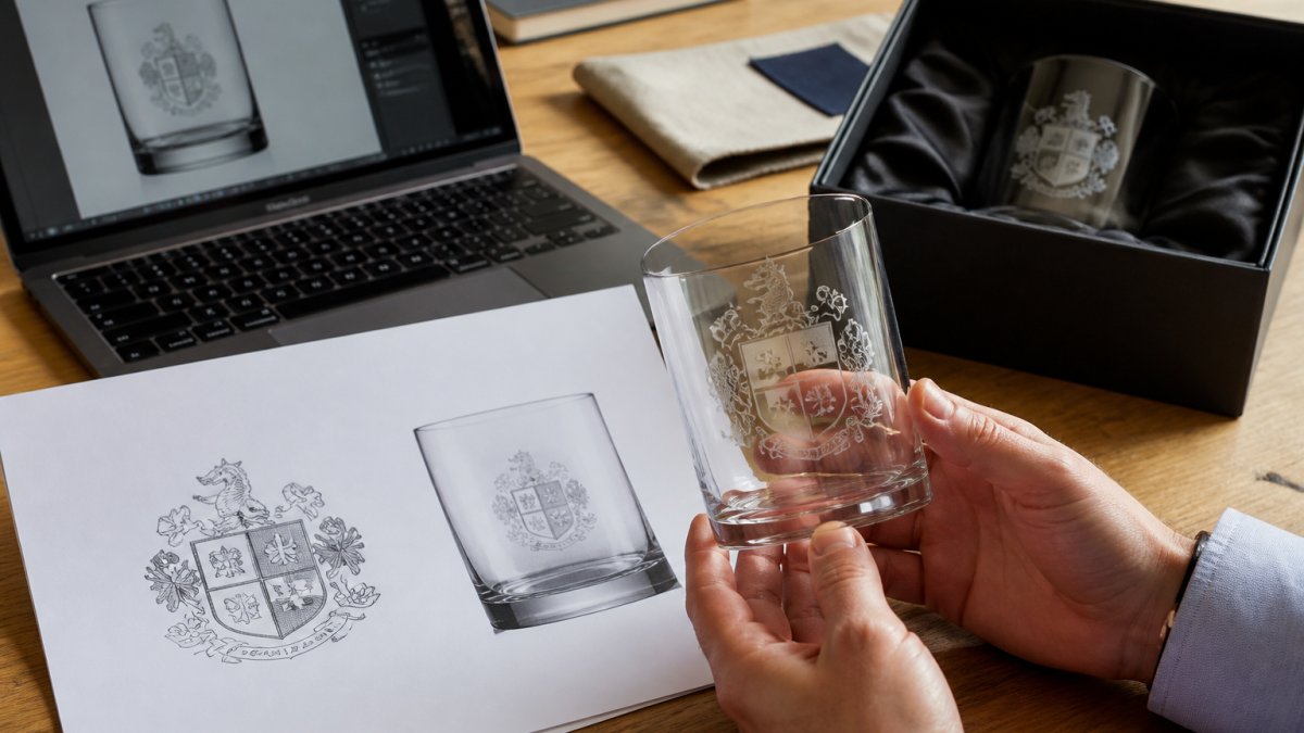 Close-up of hands holding an etched glass tumbler next to a printed artwork proof sheet on a wooden desk, with a laptop showing a logo file in the background