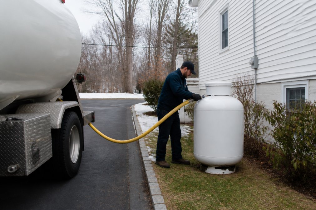 A propane delivery truck parked in the driveway of a suburban Long Island home on a grey winter morning, with a uniformed technician connecting a yellow hose to a white above-ground propane tank beside the house