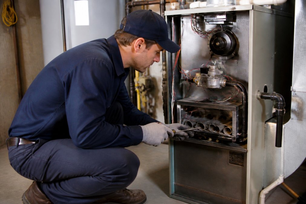 Service technician performing propane furnace maintenance, checking burner assembly and heat exchanger components with tools in home furnace room