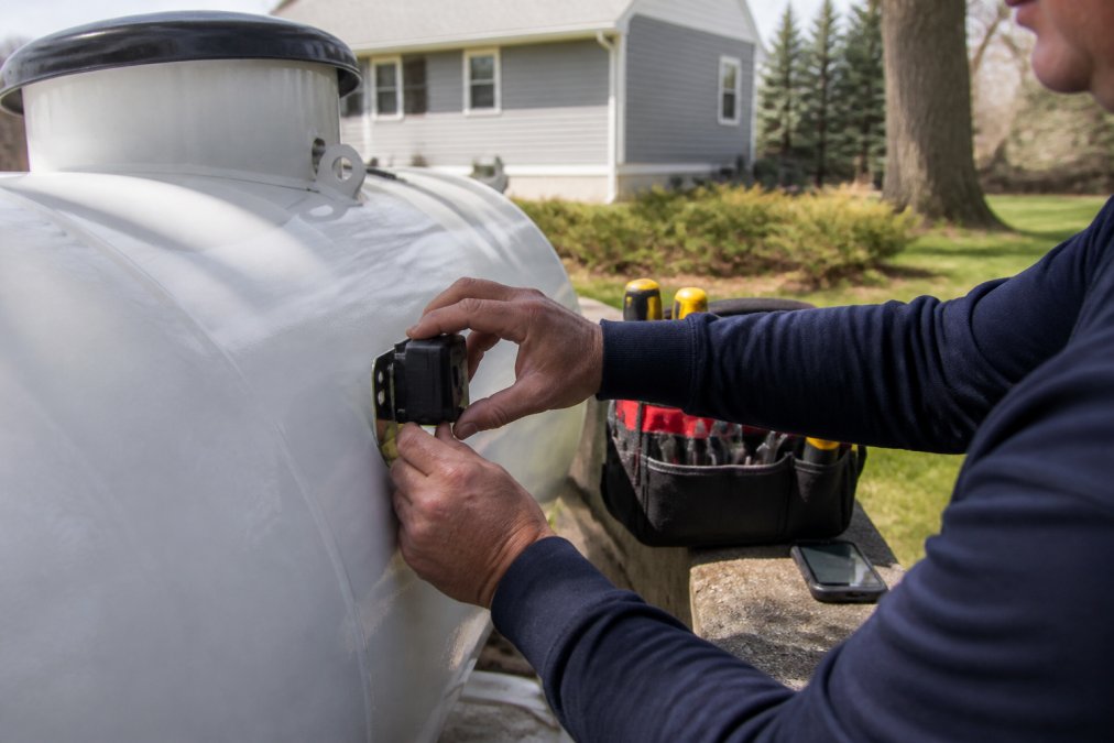 Technician's hands installing a wireless sensor device on the side of an outdoor residential propane tank, with a smartphone displaying the monitoring app visible in the background and installation tools nearby on a clear day