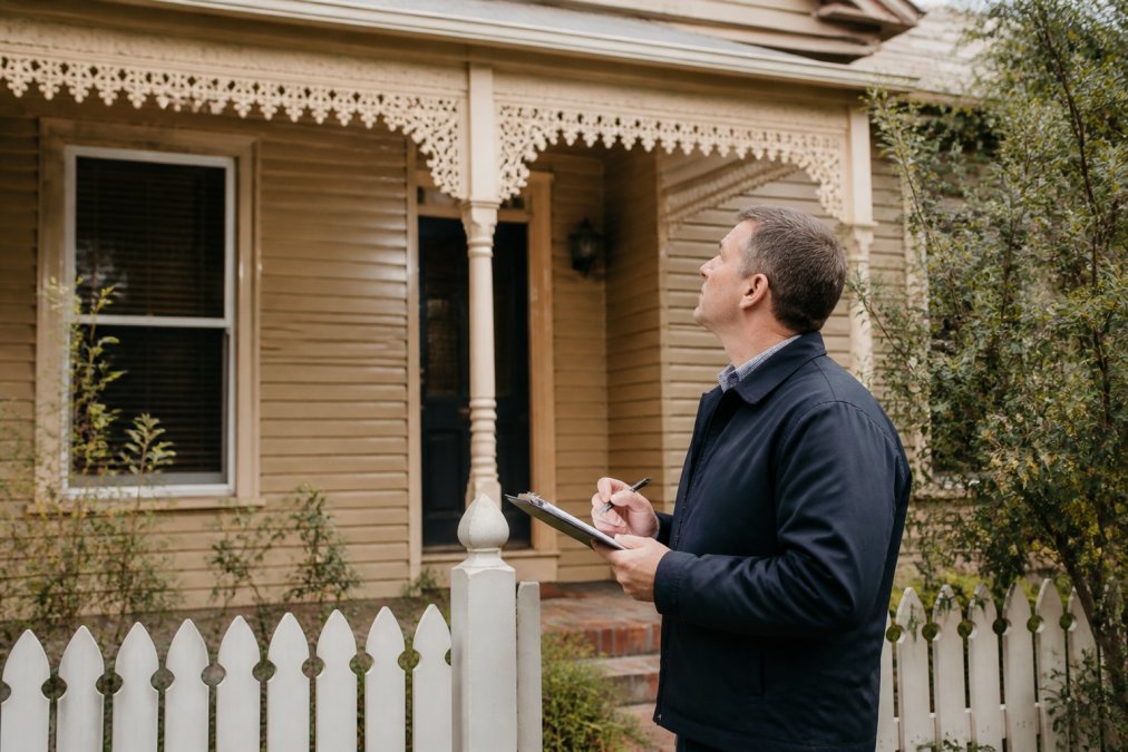 Professional accredited valuer inspecting residential property exterior with clipboard, taking measurements and notes, examining building condition and structural elements in natural daylight